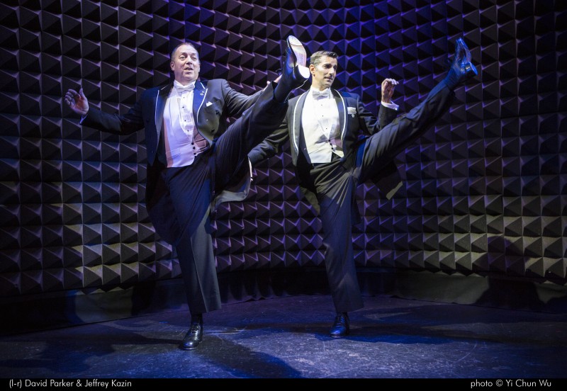 David Parker and Jeffrey Kazin wear tuxedos. Standing side by side, they kick one leg up to the audience. 